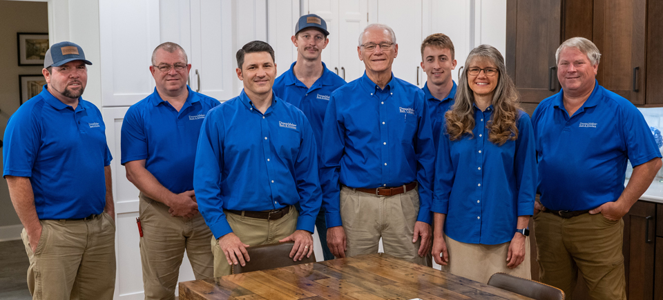 Eight people in matching blue shirts and khaki bottoms stand together in a kitchen, smiling. A wooden table is visible in front.